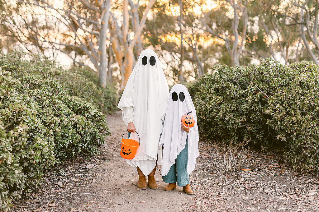 Geister-Kostüm selber machen: Zwei Kinder mit weißen Bettlaken und ausgeschnittenen Augen gehen mit Kürbiseimern auf Halloween-Tour.