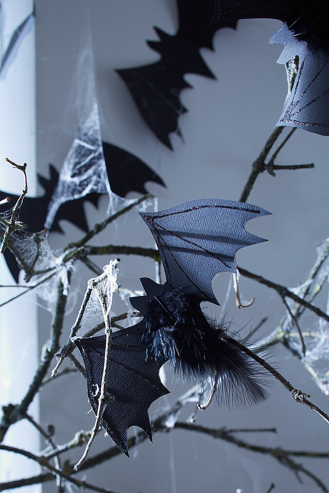 Halloween Deko selber machen: Schwarze Glitzer-Fledermaus mit Federn sitzt als gruselige Dekoration auf einem kahlen Ast.