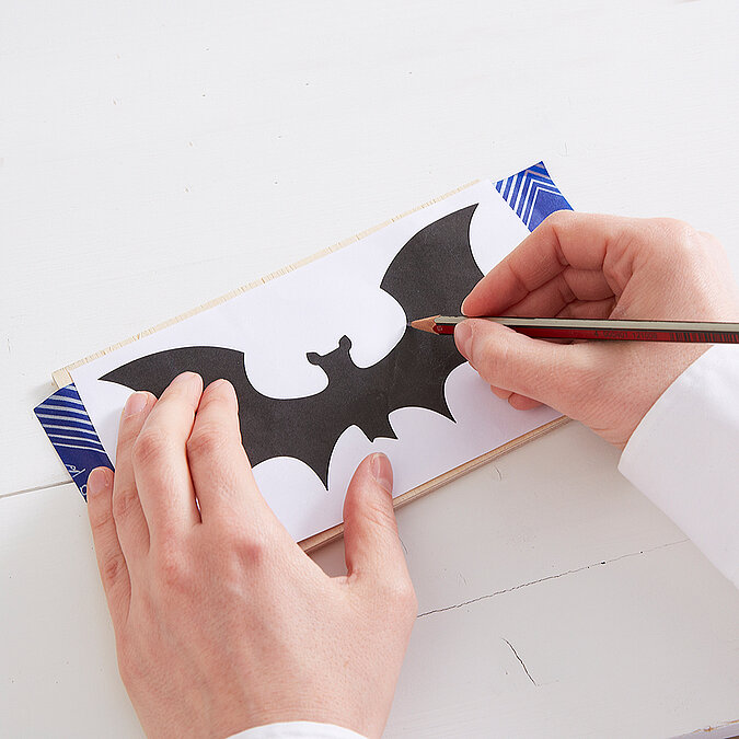 Halloween Deko selber machen: Eine Fledermaus-Vorlage wird auf Holz übertragen und für die Dekoration angezeichnet.