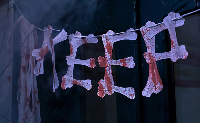 Halloween Deko selber machen: Detailansicht einer Halloween-Girlande aus Papierknochen mit gruseligem Schriftzug vor dunklem Hintergrund.
