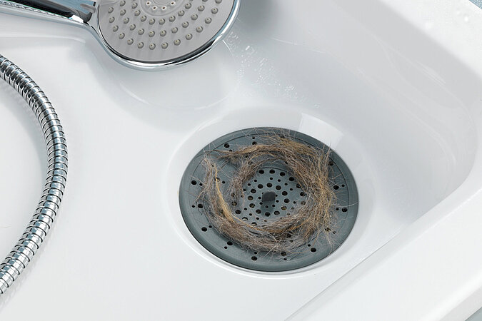 Ein Duschkopf liegt in eine Dusche daneben ein Haarsieb mit Haaren auf dem Abfluss