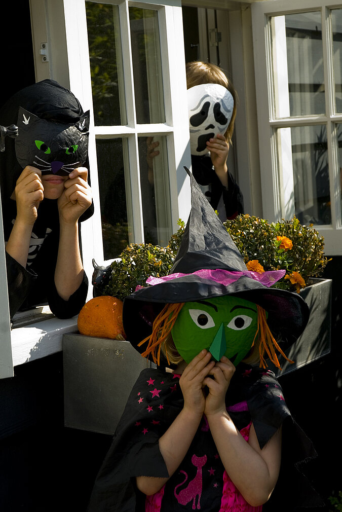 Halloween-Kostüm selber machen: Kinder präsentieren selbst gebastelte Hexenmasken aus Pappmaché in verschiedenen Farben.