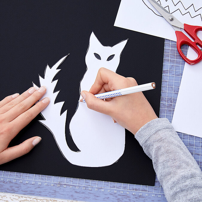 Halloween Deko selber machen: Katzenmotiv und weitere Figuren werden als Vorlage auf schwarzes Papier übertragen.