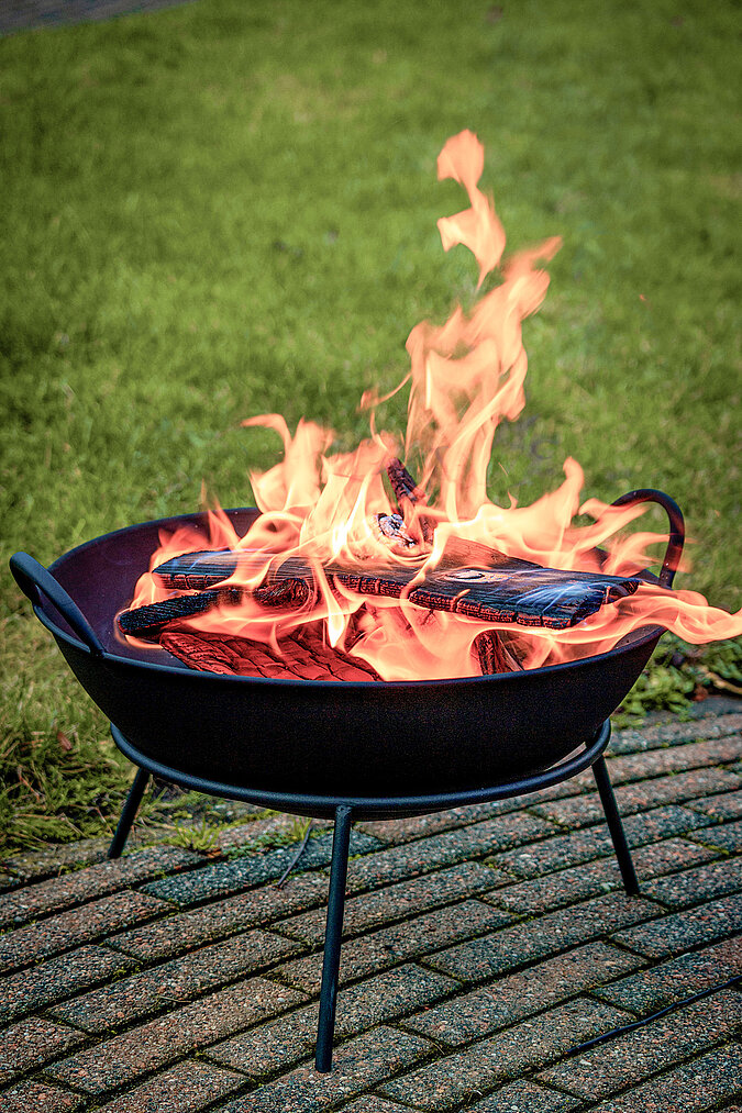 In einer Feuerschale lodern Flammen über glühendem Brennholz im Garten.