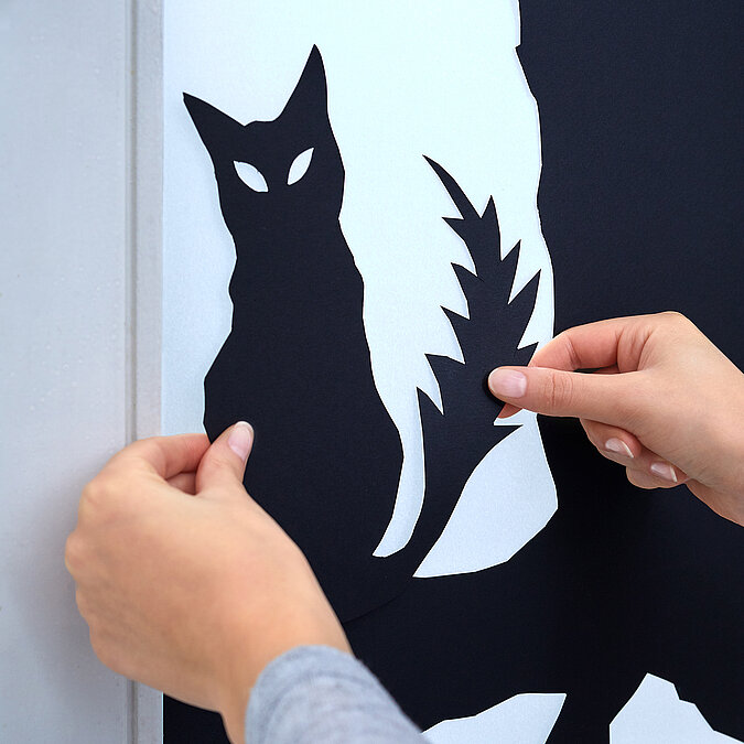 Halloween Deko selber machen: Schwarzes Katzenmotiv wird als Halloween-Fensterbild auf eine helle Fläche geklebt.