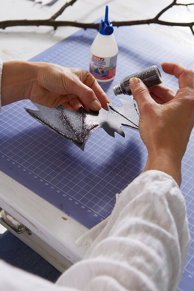 Halloween Deko selber machen: Schwarze Papierfledermaus wird mit Kleber und Glitzer für eine schaurige Dekoration verziert.