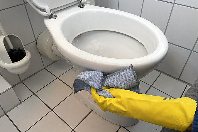 Hand mit Putzhandschuh wischt Toilette von aussen mit einem Lappen 