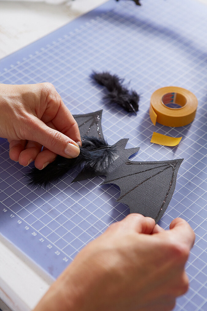 Halloween Deko selber machen: Selbst gebastelte Fledermaus wird mit schwarzen Federn und Glitzerdetails dekoriert.