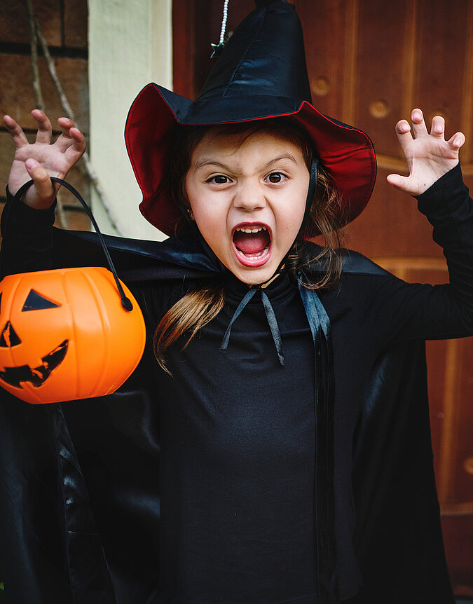 Hexen-Kostüm selber machen: Kind mit schwarzem Umhang und Hexenhut hält einen Kürbiseimer und zieht eine gruselige Halloween-Pose.