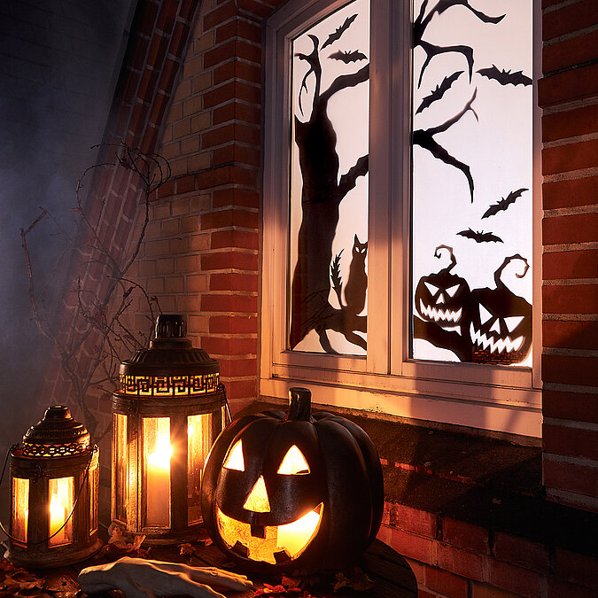 Halloween Deko selber machen: Beleuchtetes Fenster mit schwarzen Halloween-Motiven, Kürbislaternen und Kerzen vor dem Haus.