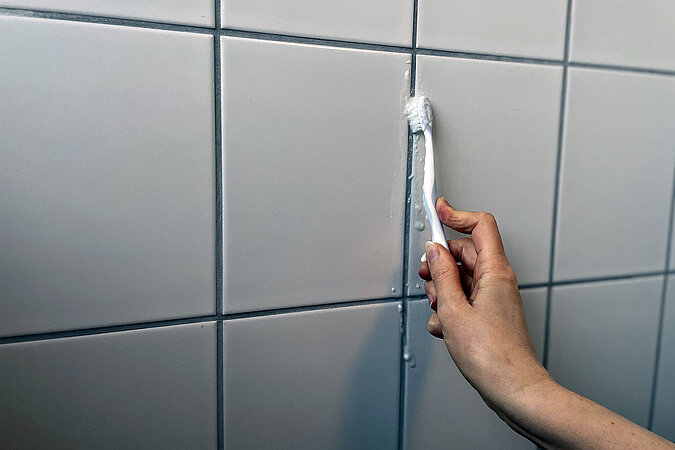 Detailaufnahme einer Hand die mit Zahnbuerste und Hausmittel die Fliesenfugen reinigtugen 