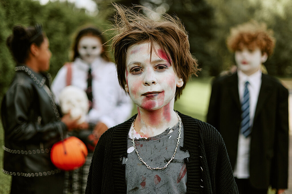 Halloween-Kostüm selber machen: Kind mit weiß geschminktem Gesicht und zerzaustem Haar im Zombie-Kostüm, im Hintergrund weitere verkleidete Kinder.